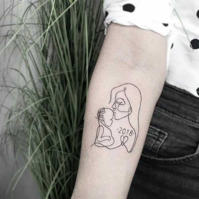 Tatouage minimaliste sur l'avant-bras d'une mère embrassant son bébé avec l'année de sa naissance